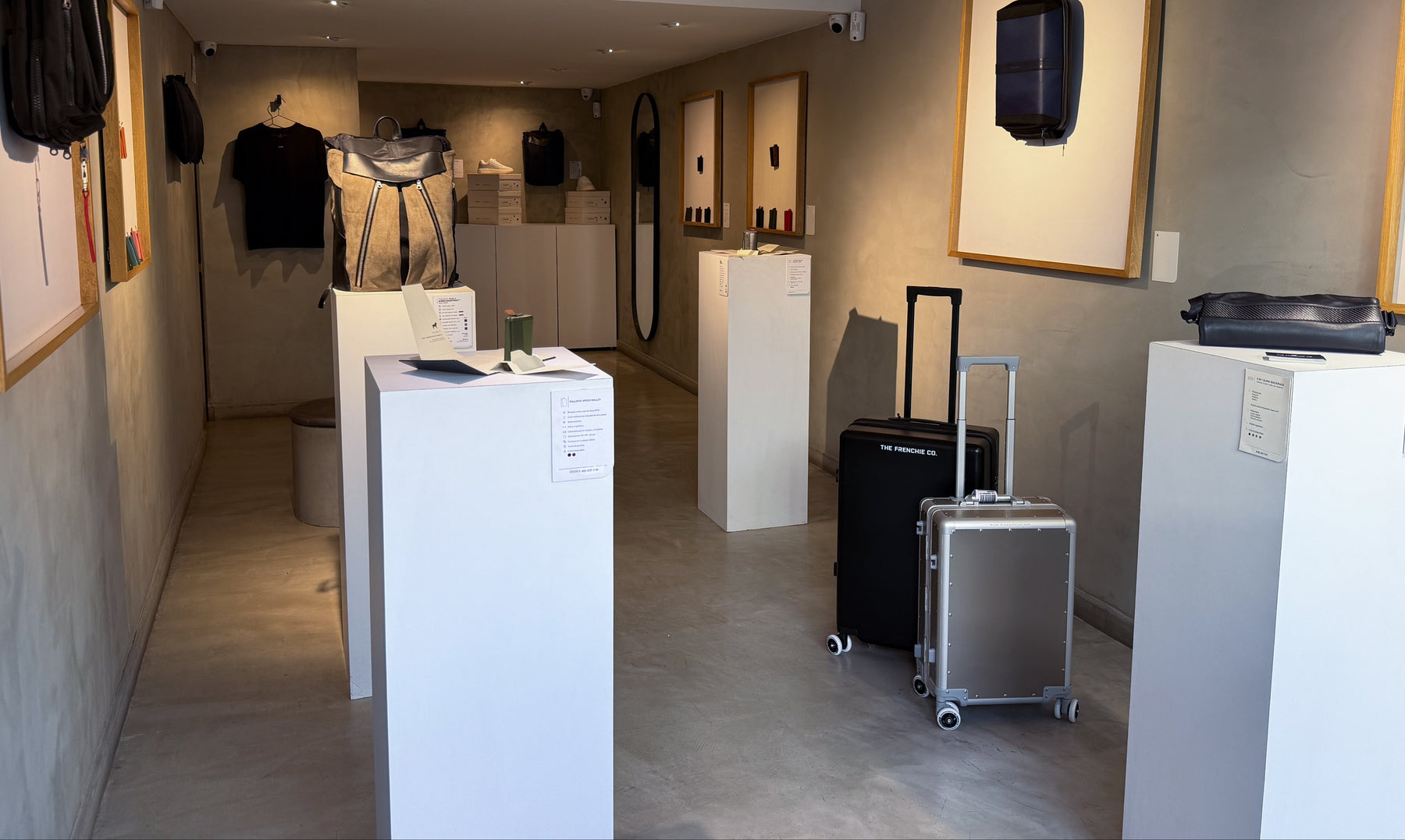 A retail showroom interior with white display pedestals, framed wall art, and two rolling suitcases on the floor. The sign 'THE FRENCHIE CO.